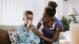 A boy blows his nose into a tissue, his mother comforting him during his illness.