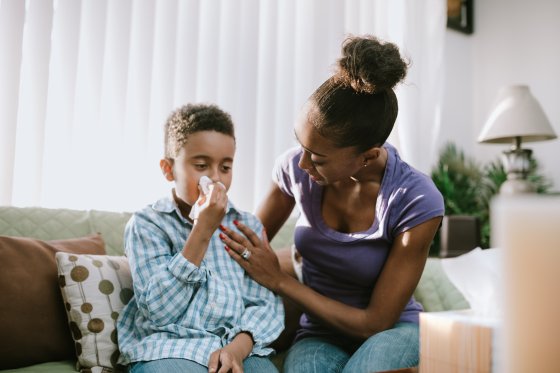 A boy blows his nose into a tissue, his mother comforting him during his illness.