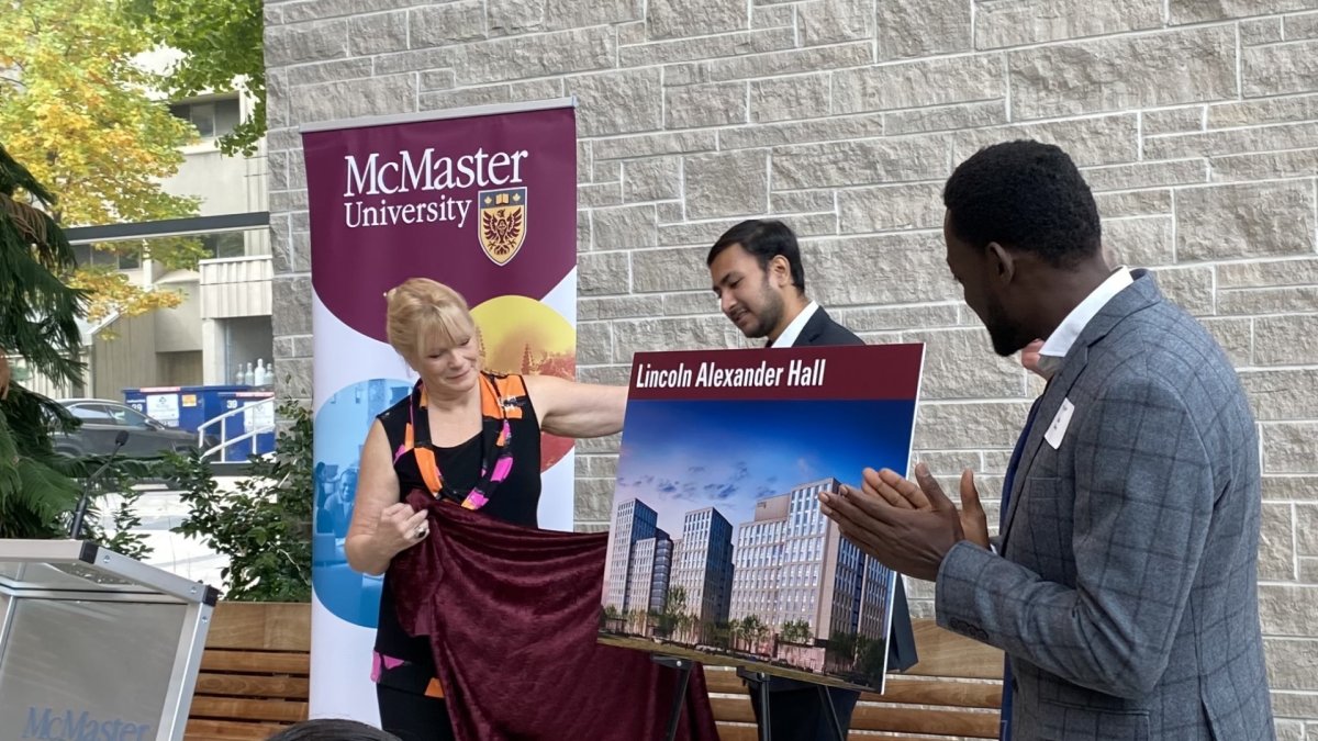 A woman holds a curtain after unveiling a design photo of McMaster's newly planned student residence, while two men also stand next to the image - one of the men is applauding.