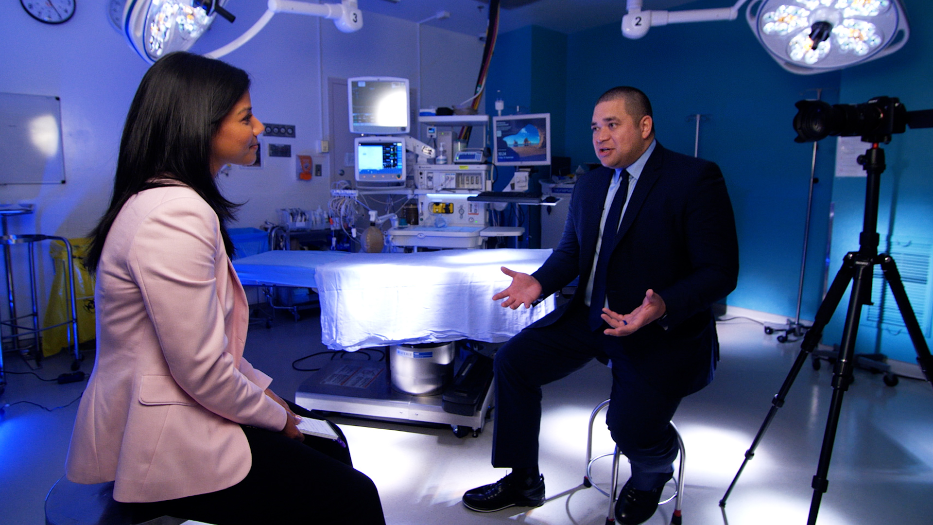 Neetu Garcha is wearing a light pink blazer in an operating room where she is interviewing Dr. Alika Lafontaine. They are engaged in