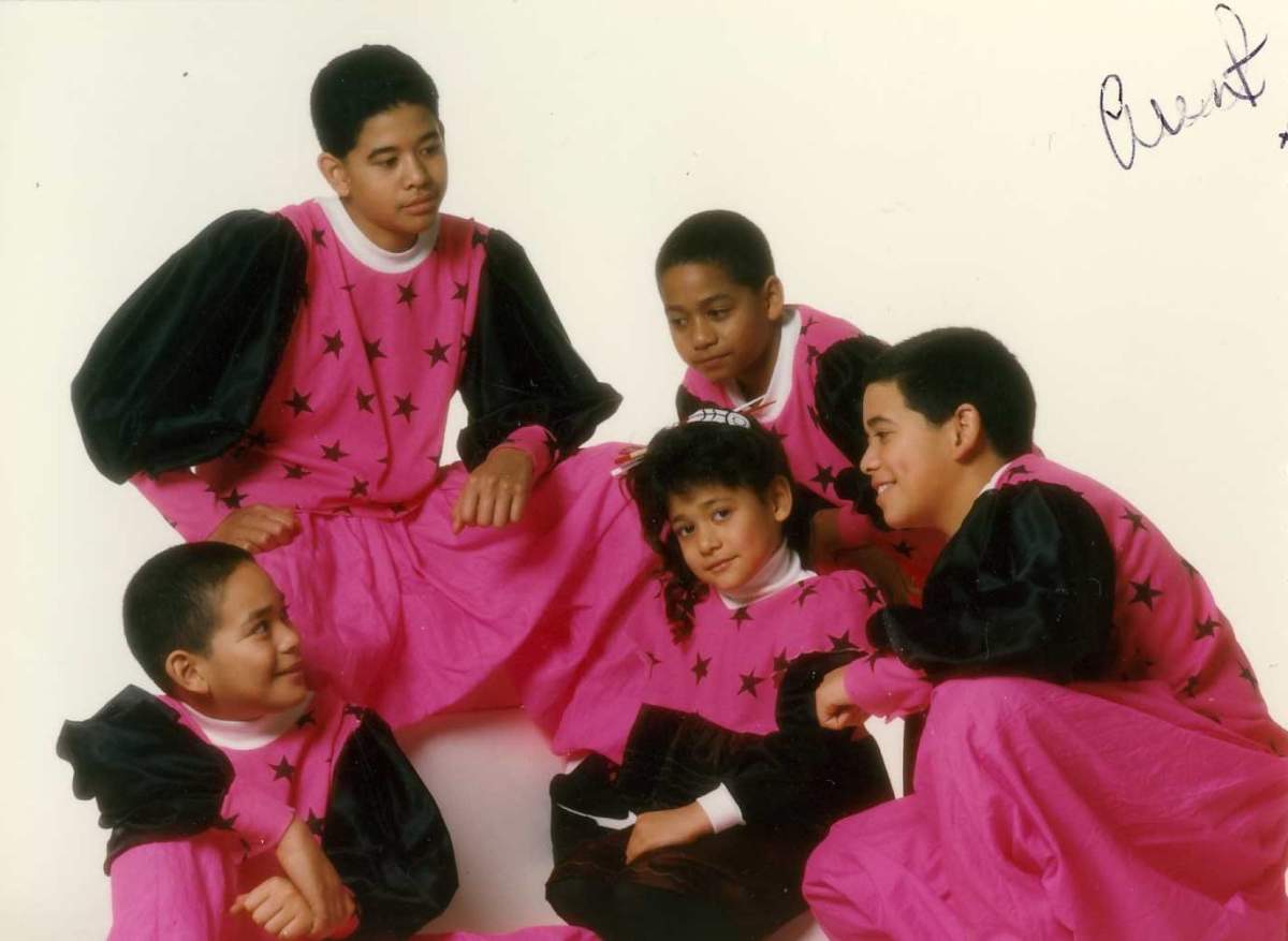 A young pop-group, dressed in matching pink and black ensembles, sits together for a photo. The one young girl in the photo, looks towards the camera.