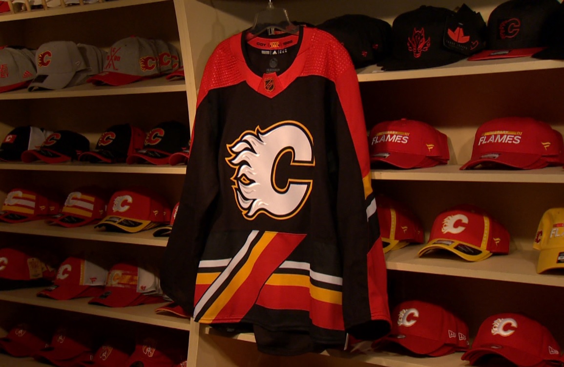 The Calgary Flames Reverse Retro jersey hangs in front of a display of hats. The jersey has a red, orange, black and white sash with a diagonal pedestal supporting a white 'Flaming C'. The shoulders and arms feature red piping and a retro NHL crest is fixed at the neck of the jersey.