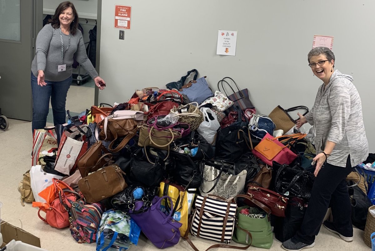 Two women pose on either side of a pile of purses of various colours, shapes and sizes.
