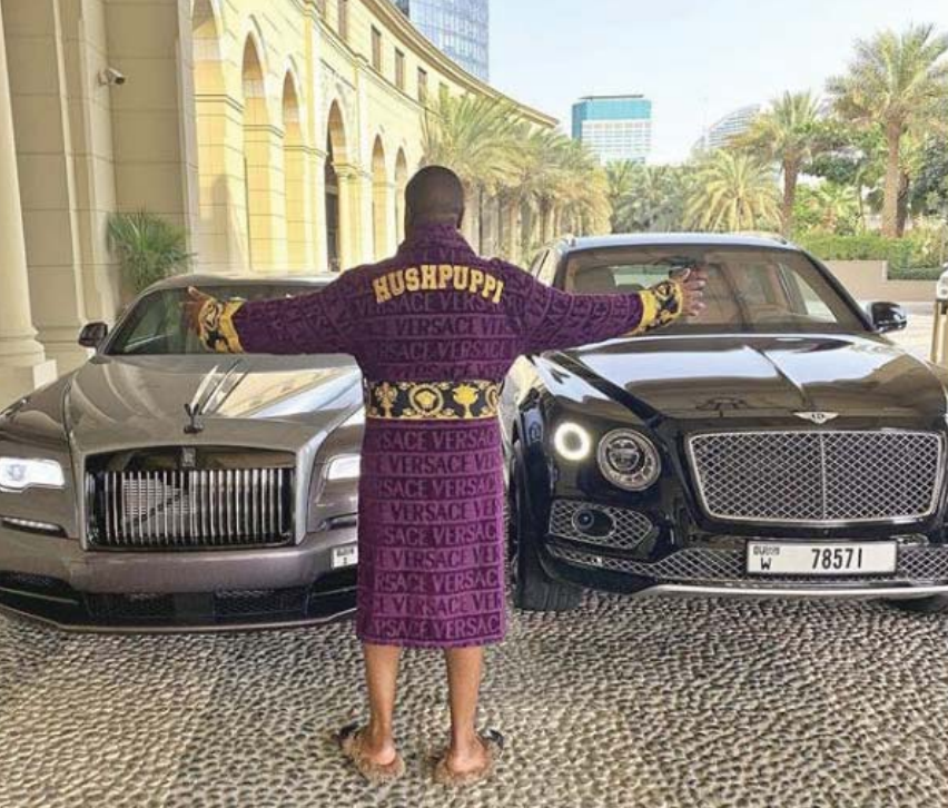 Ray Hushpuppi stands in front of two cars wearing a Versace robe and slippers.