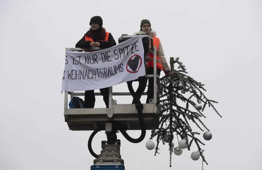 Two protesters stand on a hydraulic lift. They stand in front of a banner. One is holding the sawed-off Christmas tree tip.