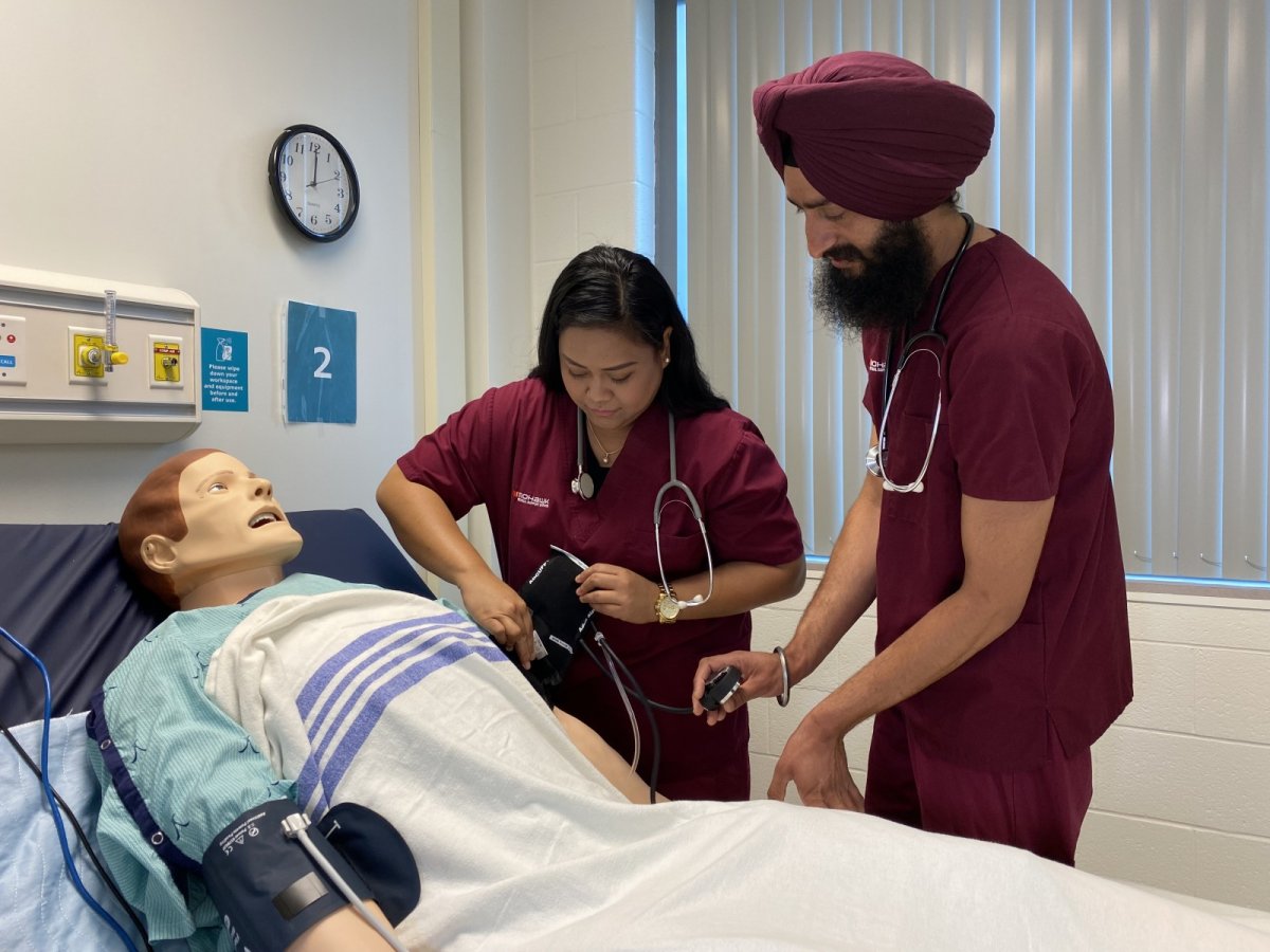 Two students from Mohawk College's PSW program work with a dummy in a simulated care room as part of their training.