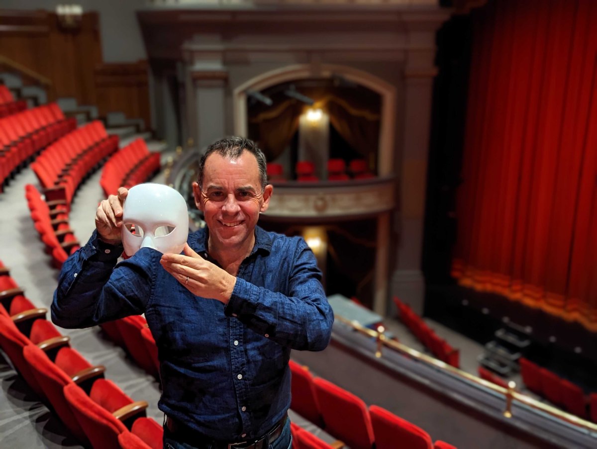 A man, Dennis Garnhum, poses wearing a blue shirt and holding a white mask in a stage at the Grand Theatre.