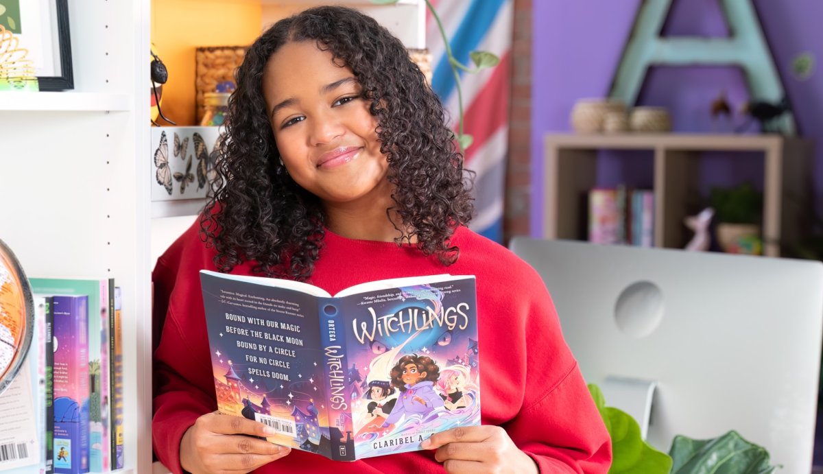 Ainara Alleyne smiles while looking at the camera and holding a copy of Witchlings, a novel by Claribel A. Ortega.