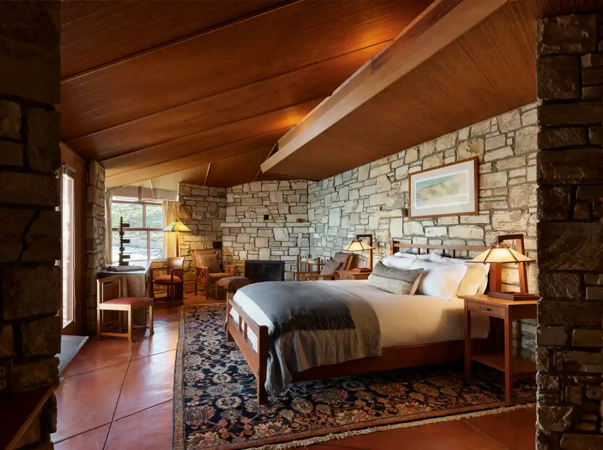 The bedroom. There is a bed and windows. The wall is stone. The flooring is wooden.