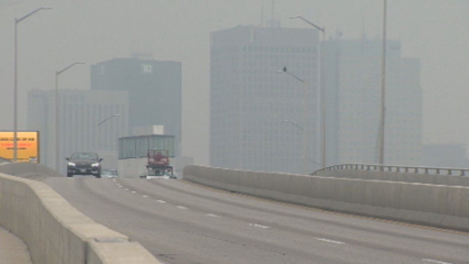 Downtown Winnipeg buildings are partially blurred by smoke in the air. A car crosses a bridge in the foreground.