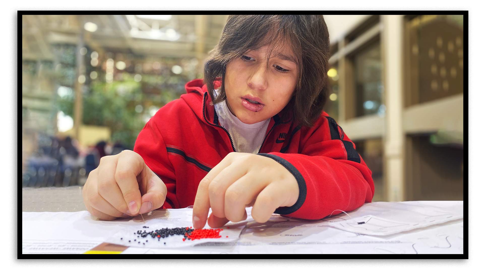 A 13-year-old boy sits at a table, learning beading techniques. He has long brown hair and wears a red sweater with black stripes down the sleeves and around the cuffs. He holds a needle in his right hand and concentrates as he tries to thread the needle through small black and red beads that are on a piece of paper. He’s in the two-storey atrium of a hotel’s event space and some indoor plants can be seen over his right shoulder.