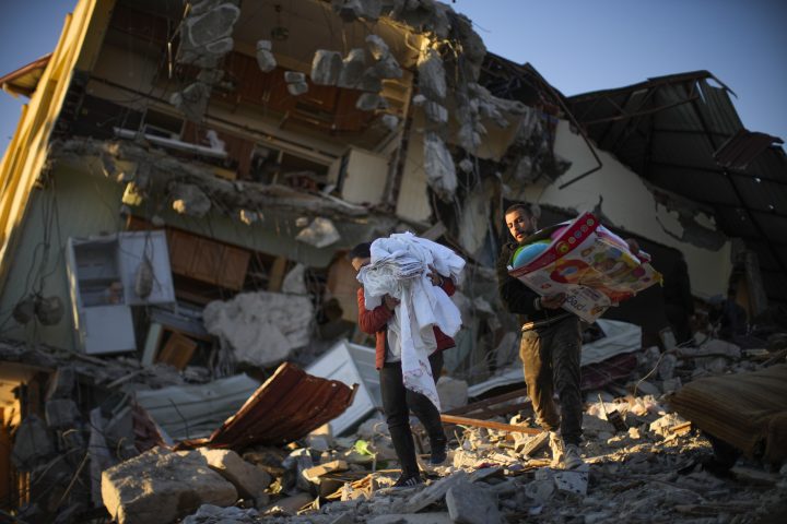Two people carry personal items as they walk past destroyed structures