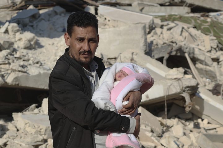 A man holds a newborn baby wrapped in a pink blanket with a destroyed building behind them