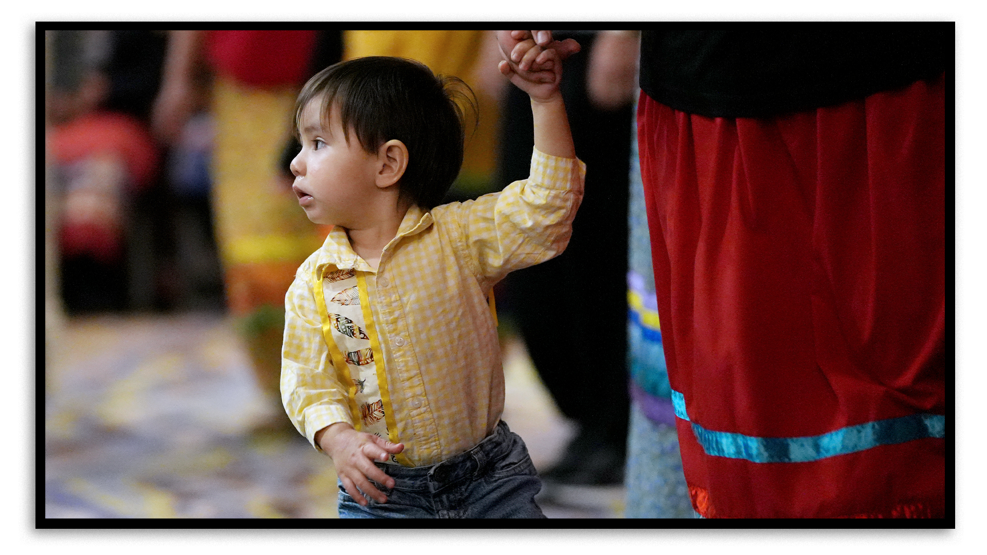 A young boy with brown hair and wearing a yellow shirt and jeans holds an adult’s hand during a round dance in a hotel event space. The woman holding his hand is wearing a red dress with a sky-blue ribbon stripe. Other attendees walk by in the background.