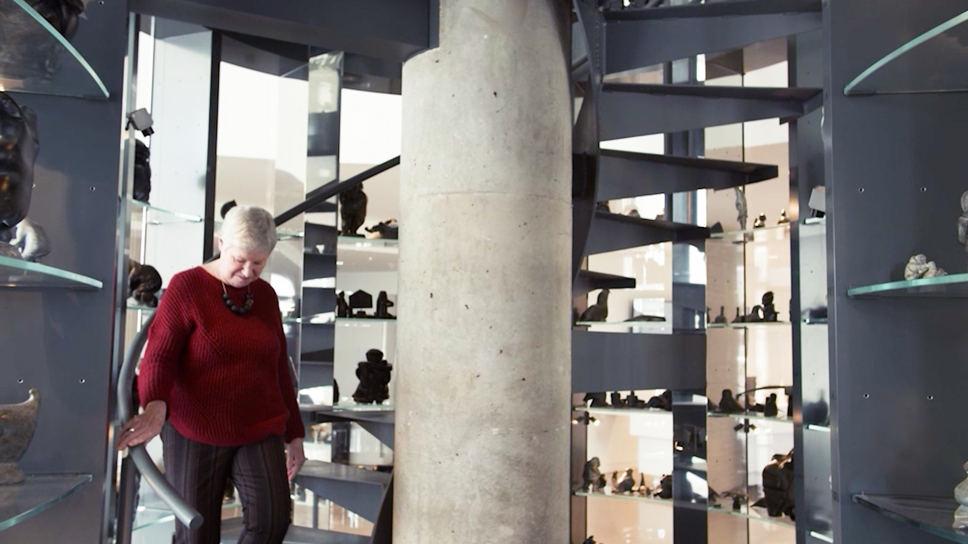 Darlene Coward Wight, Inuit Art Curator at Quamajuq-Winnipeg Art Gallery, walks down the stairs inside the visible vault. She's wearing a dark red sweater and dark grey paints. She has cropped white hair and is looking down.