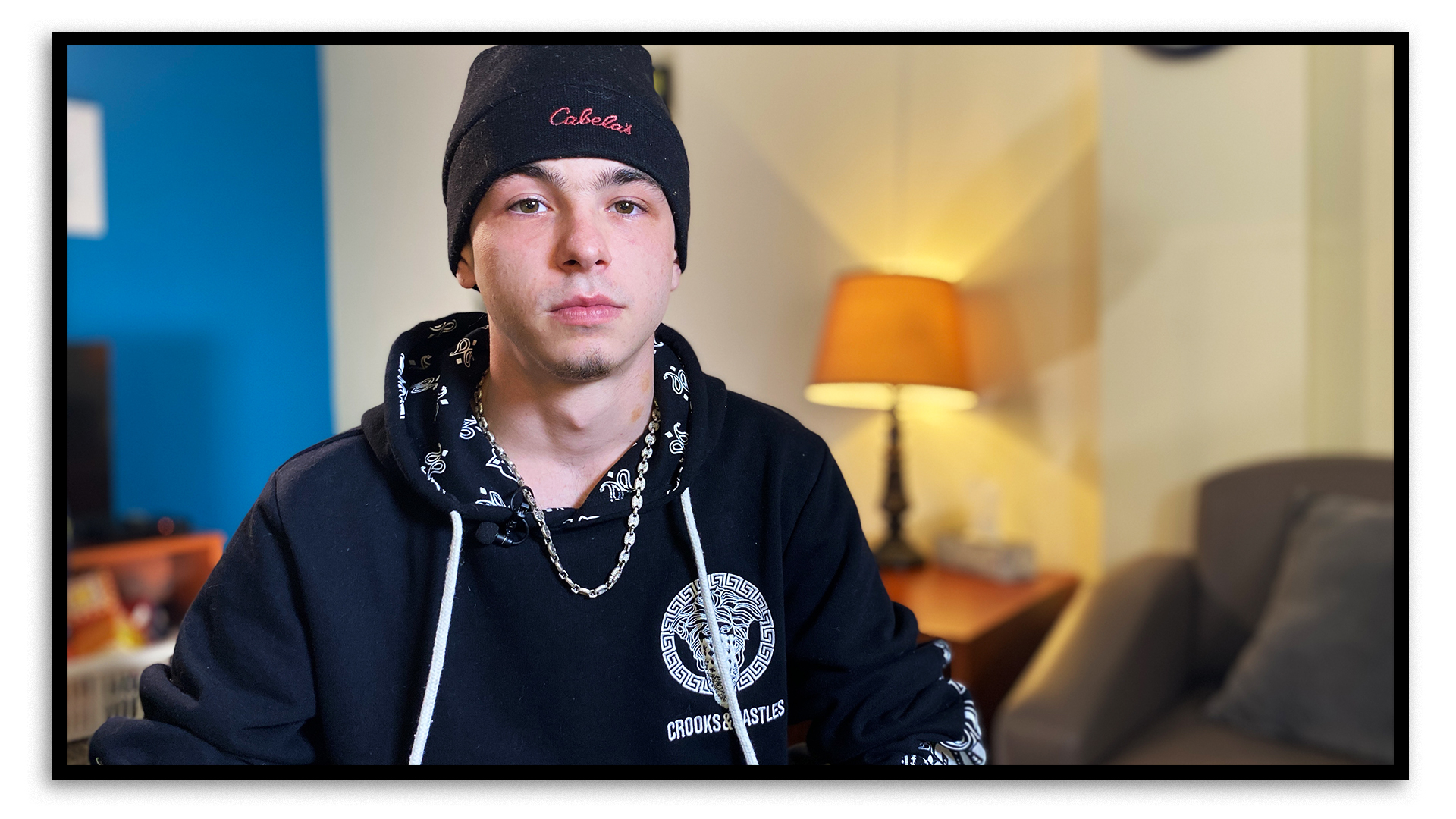 Ethan Pokno, aged 21, sits in a conference room at Nogdawindamin's offices. He's wearing a black sweatshirt with a white logo on the left side of his chest, a necklace and a black toque. Behind him, there’s an armchair and a table tamp that gives off a warm glow.