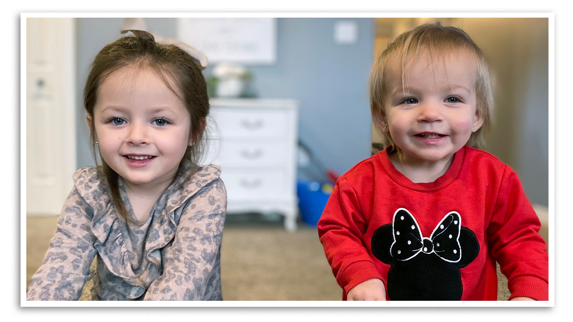 Two little girls smile for a photo in a playroom. Blue-eyed Grace, aged 4, is on the left, wearing a beige coloured shirt with darker spots and with a bow in her shoulder-length brown hair. She looks directly into the camera. On the right, blond-haired Hope, aged 1, wears a red sweater with a cartoon figure, Minnie Mouse, on the front.