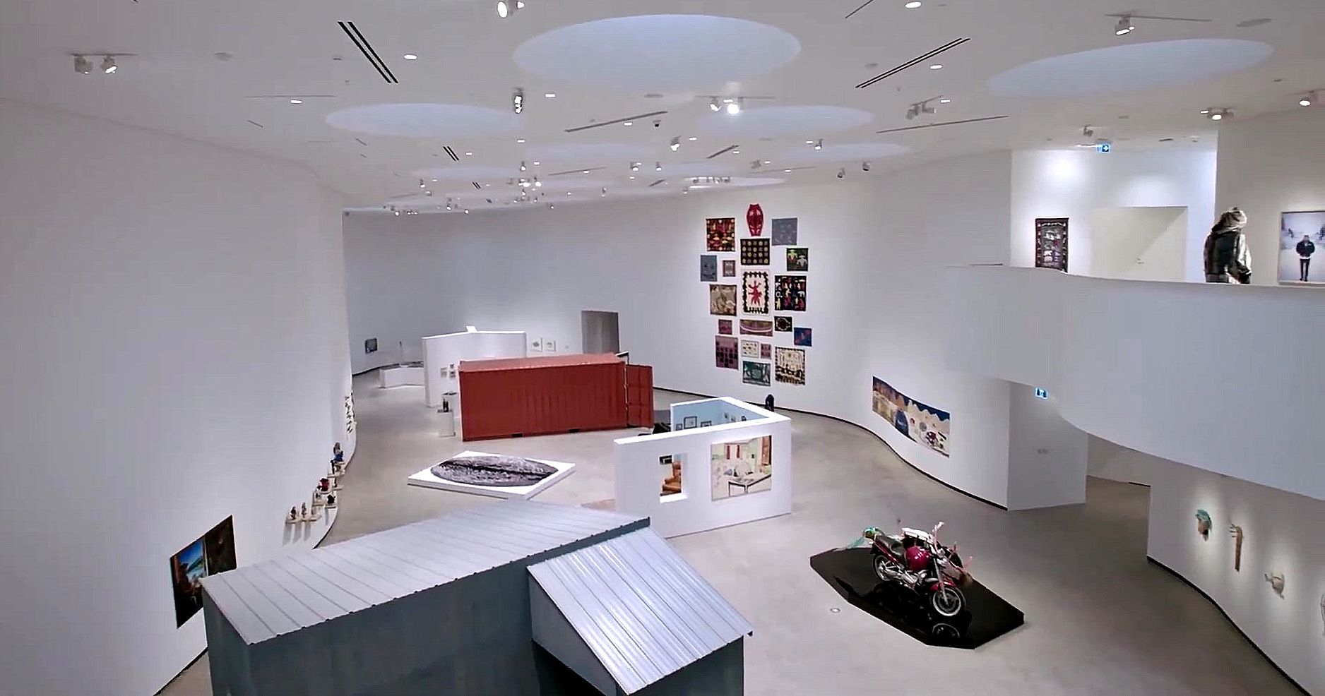 A full-size, red shipping container, doors open, is placed in the centre of a bright white exhibition room at an art gallery in downtown Winnipeg.