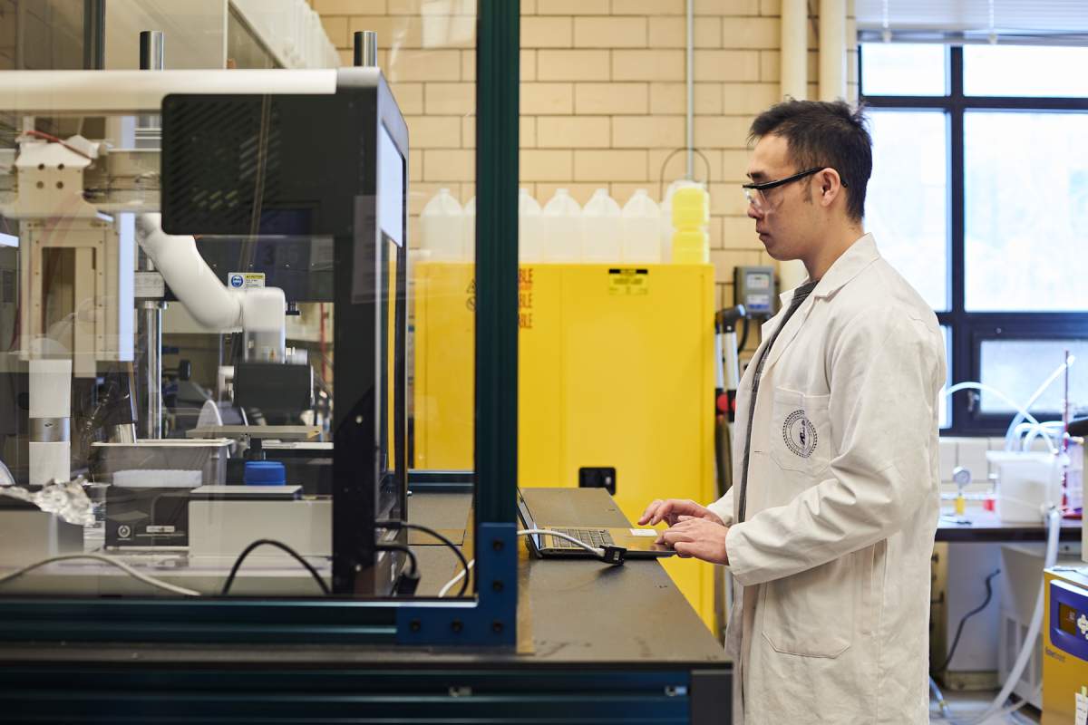 A scientist controls the robotic arm via the laptop to transfer a sample into a testing instrument.