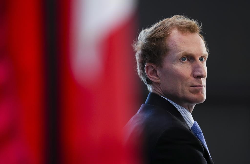 A man sits behind a Canada flag, he has short read hair and a concerned look on his face.