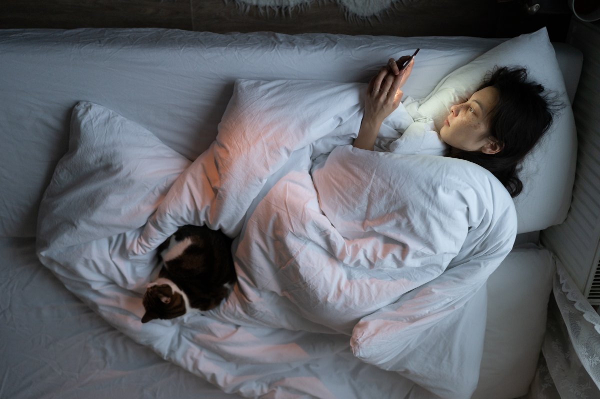A woman in bed with her cat. She is on her phone.