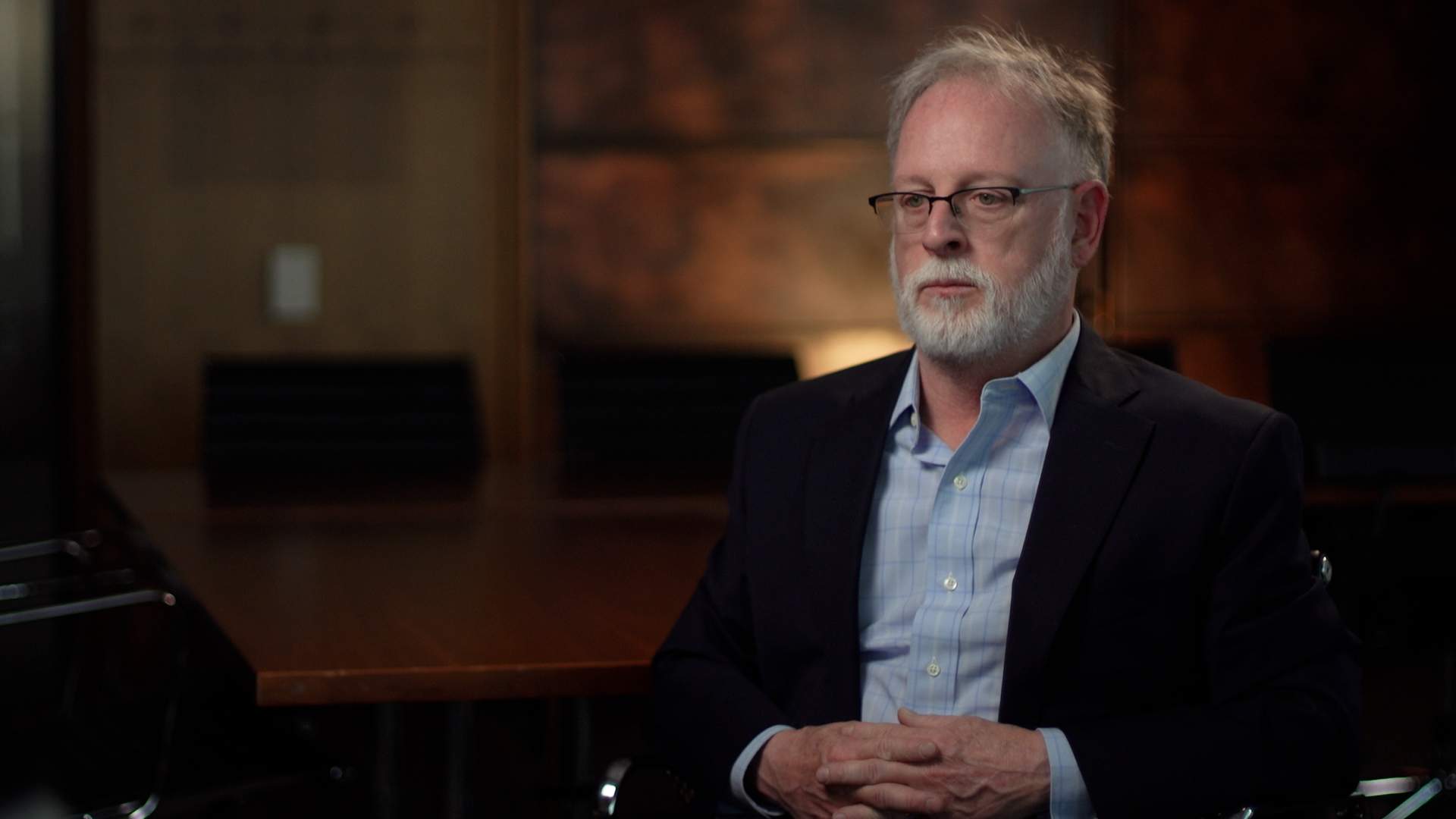 Bob Kirk, a man with a white beard and white hear, is looking to the left of the screen. He is wearing glasses, a blue collared shirt, and a blazer.