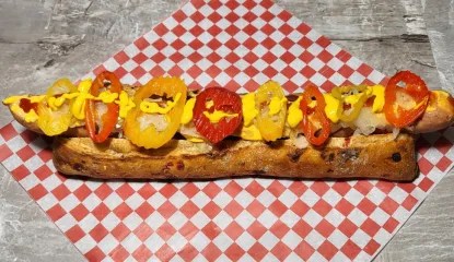 A 12-inch jalapeno cheddar dog made with aged wagyu & elk meat which has been marinated in a 2010 Lokoya Mount Veeder Cabernet Sauvignon for five days. It is served atop a bed of thinly sliced Bundnerfleisch in a red pepper and sun-dried tomato ciabatta topped with melted cheese, German sauerkraut, onions and banana peppers