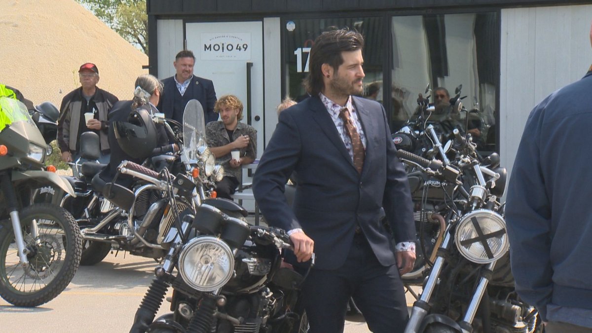 a man in a suit stands beside a motorcycle. In the background are more men in suits and motorcycles.
