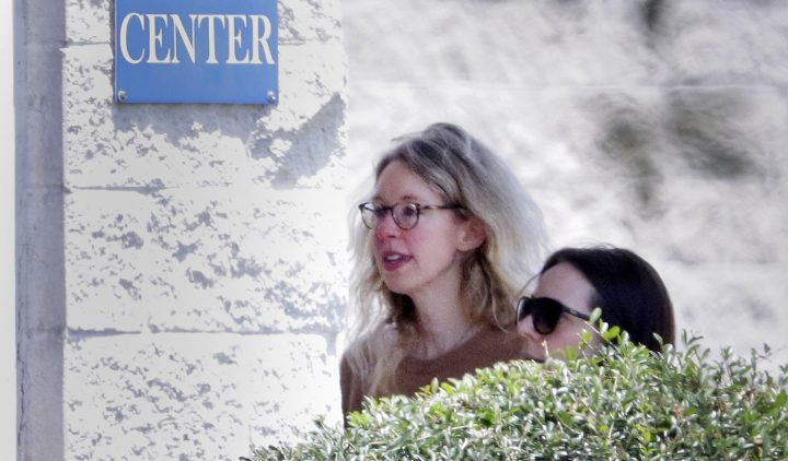 Elizabeth Holmes, wearing glasses with a light coloured shirt, walks into a building on a sunny day