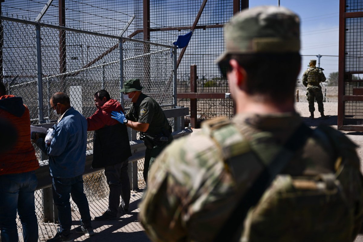 Migrants at a border crossing. There is an armed official in uniform.