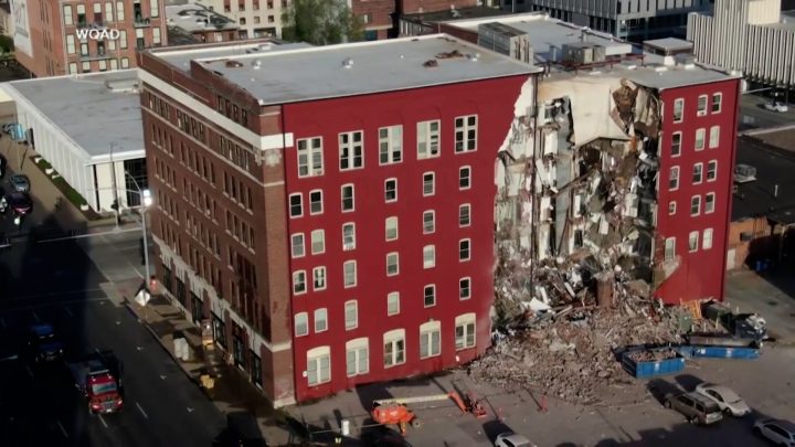 A multi-story apartment building sits with a section missing, exposing units inside, following a partial collapse of the structure