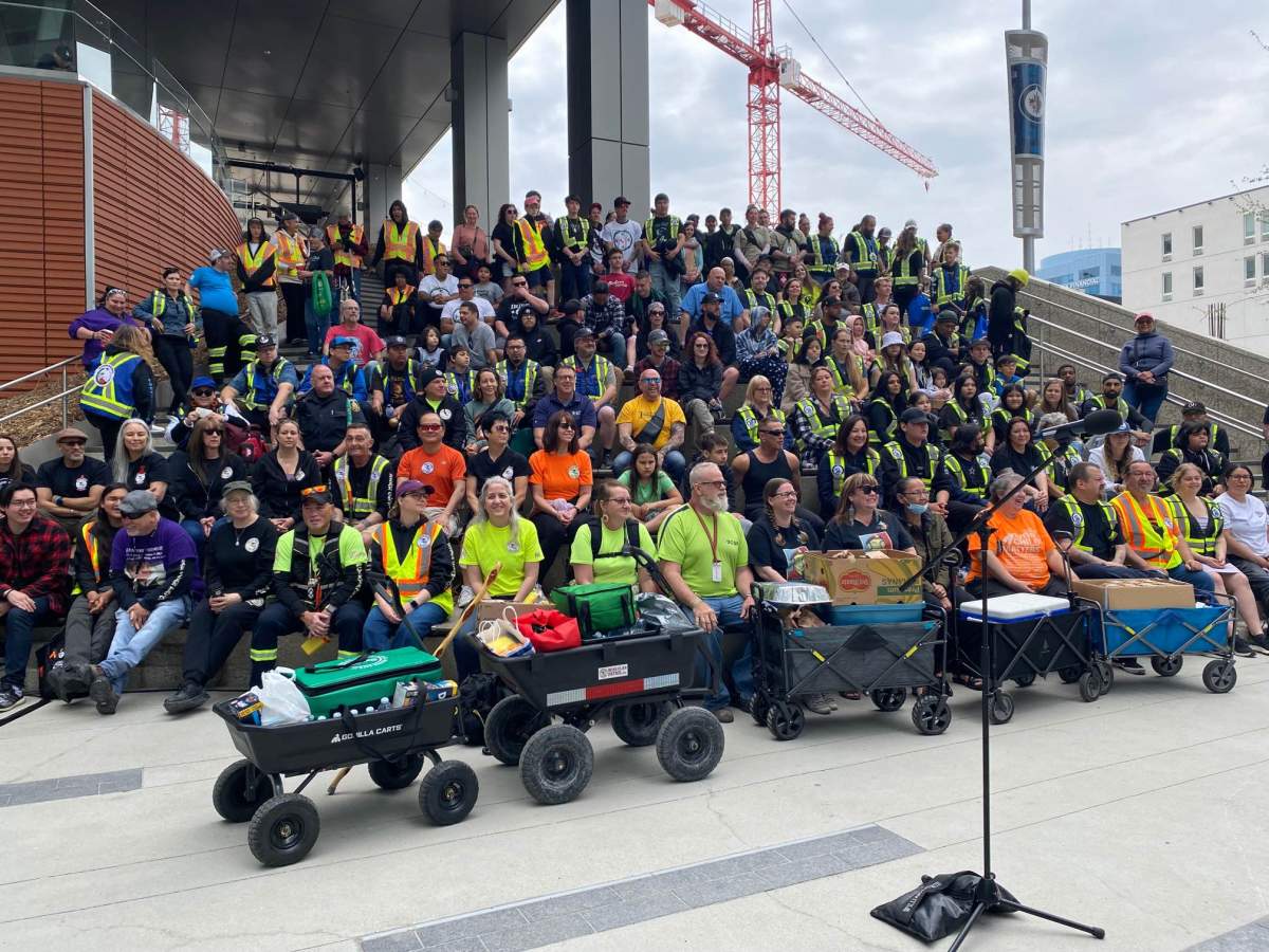 A group of people, many wearing high visibility vests, sit on a wide public staircase. Wagons full of food and supplies are in the foreground.