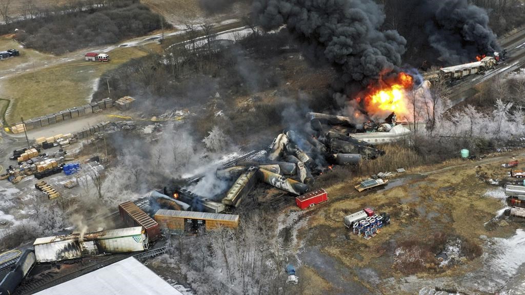 A derailed train lays among black smoke and flames.