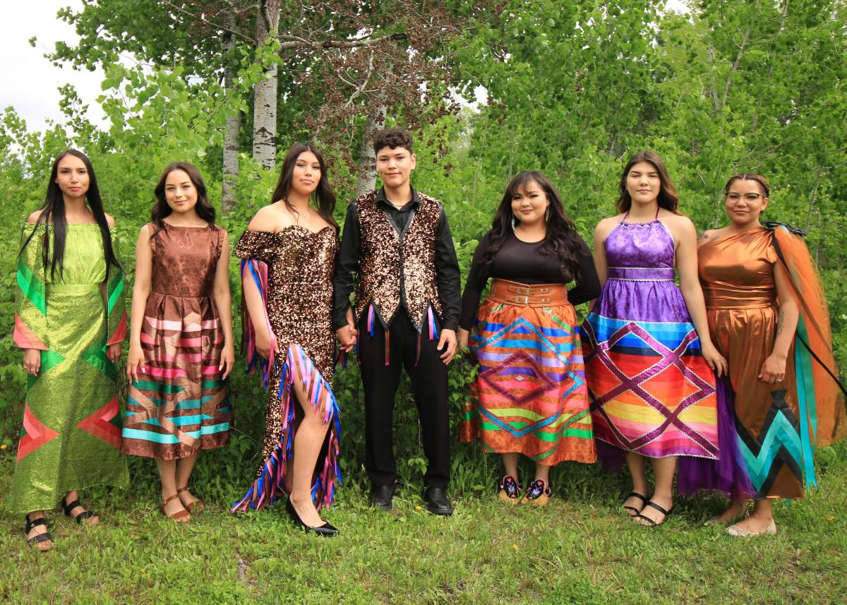 A group of people wearing ribbon skirts, shirts, stand in a line in a forested area.