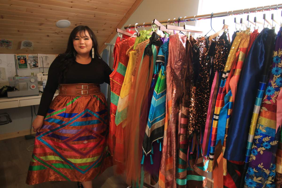 A woman in a ribbon skirt stands next to a rack of ribbon skirts.