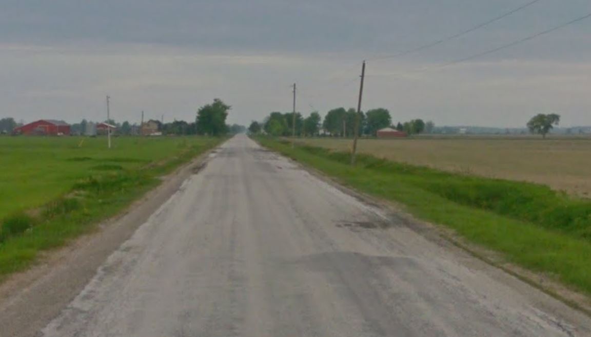 A rural road at dusk with a grassy ditch on the sides of the road and a field to the right.