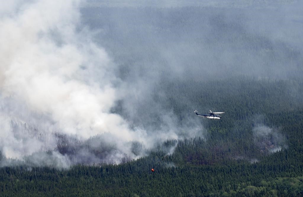 A helicopter carrying a water basket flies past a smoke plume near Lebel-sur-Quévillon, Que., Wednesday, July 5, 2023. 