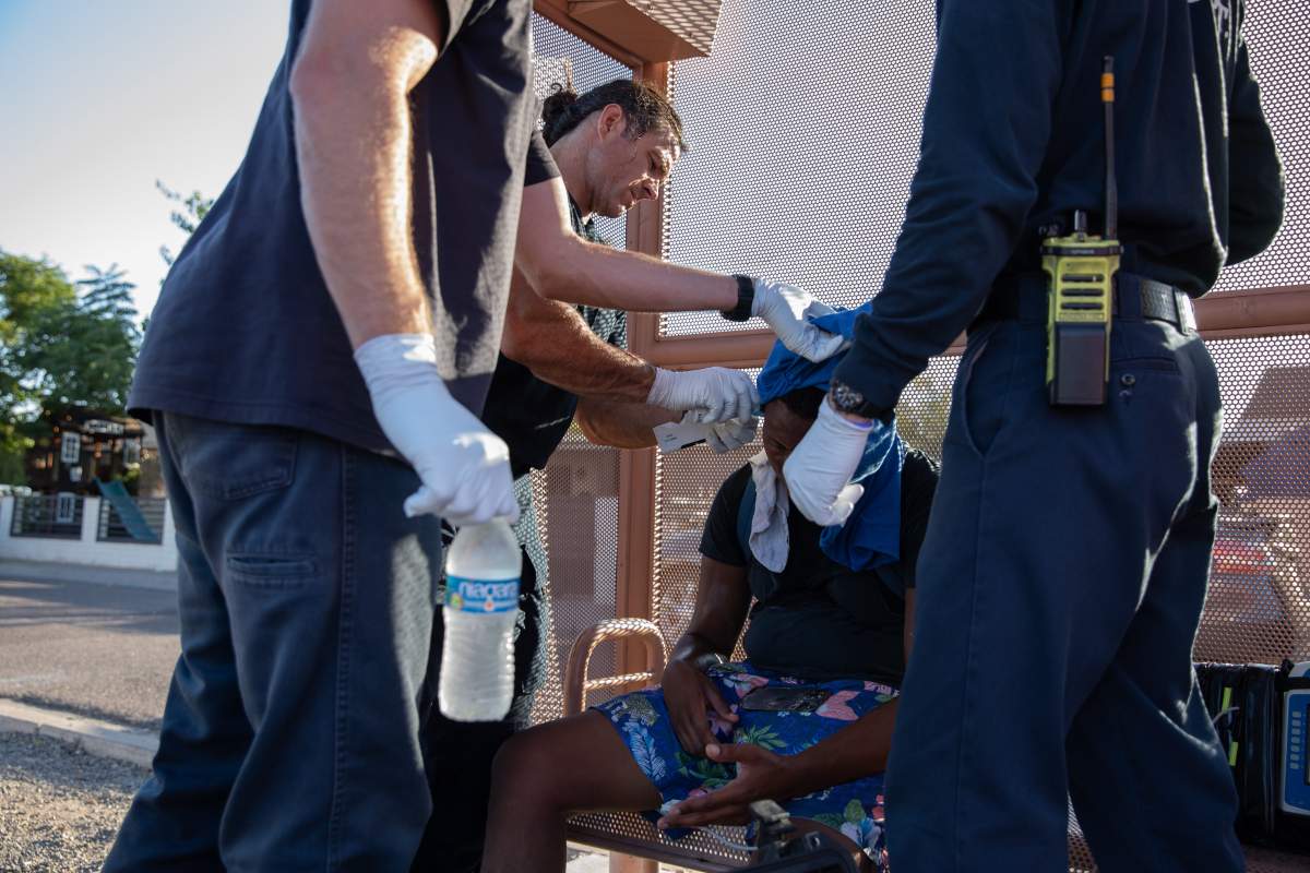 Firefighters check a man's temperature while he sits on a bus bench. One firefighter is holding a cold bottle of water.