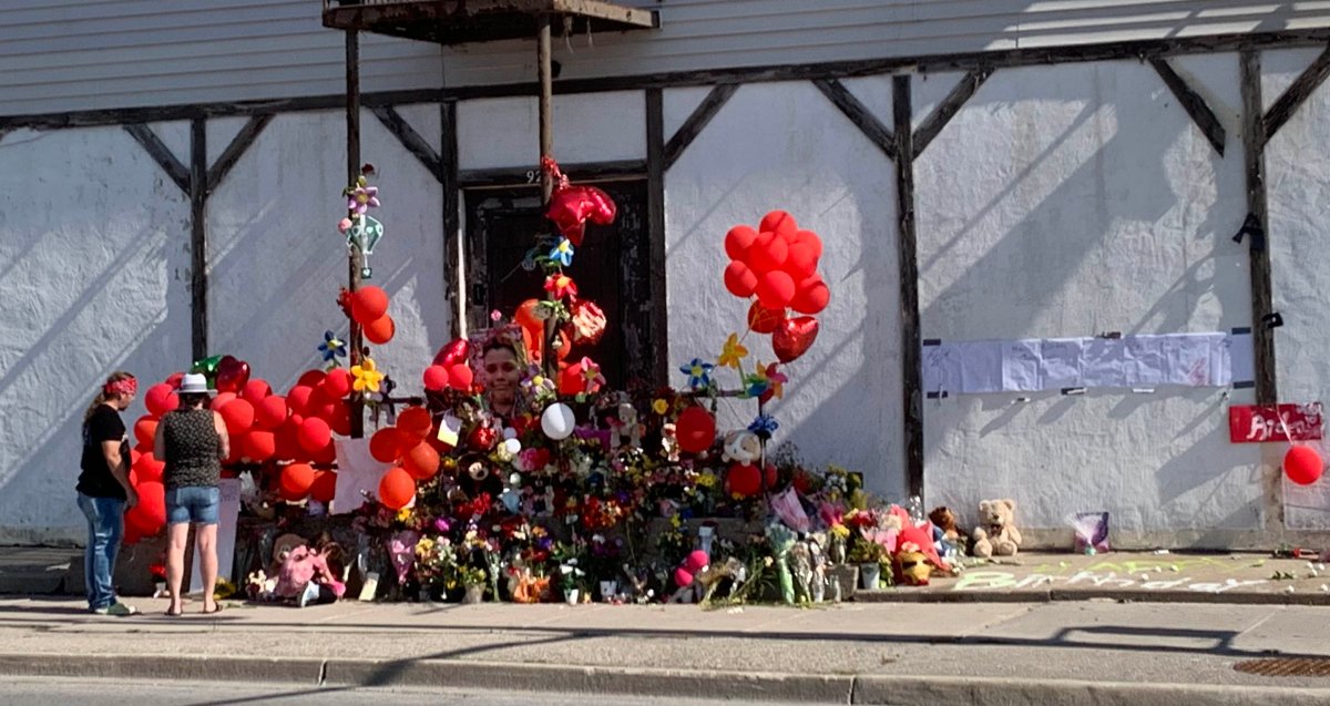 Red balloons, stuff animals, cards and other mementos rest along a sidewalk next to a white building.