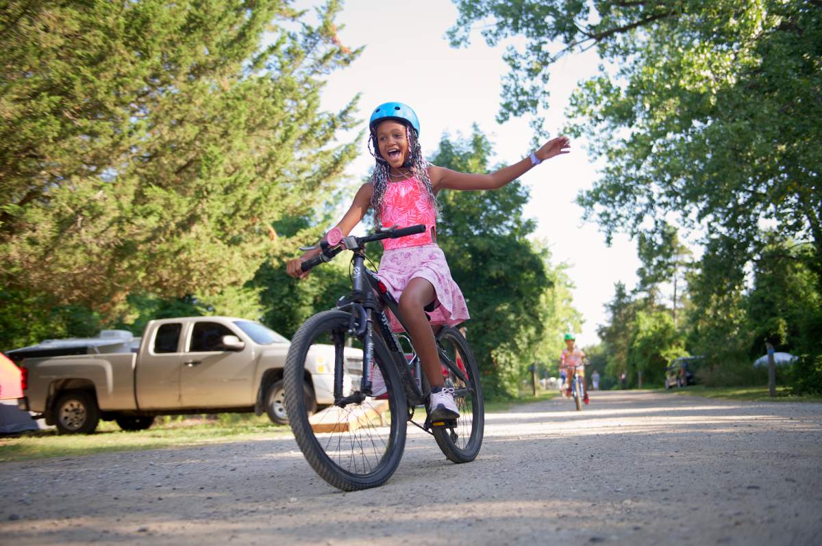 A little girl rides a bicycle on a gravel path between trees.