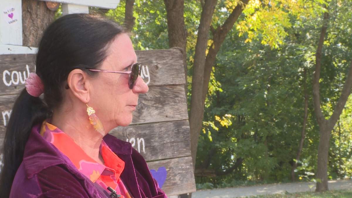 A side profile of a woman with black hair and sunglasses. Behind her are trees and a wooden sign with white lettering.