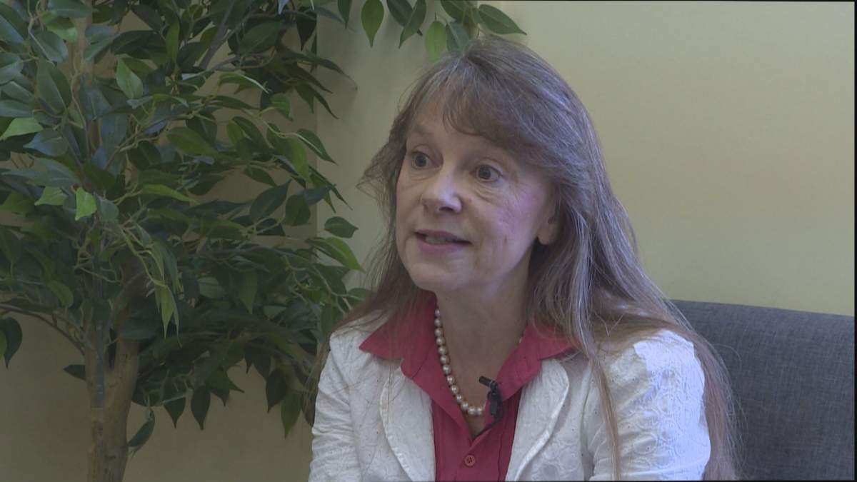 A woman wearing a white jacket and long brown hair sits in an office by a tree.