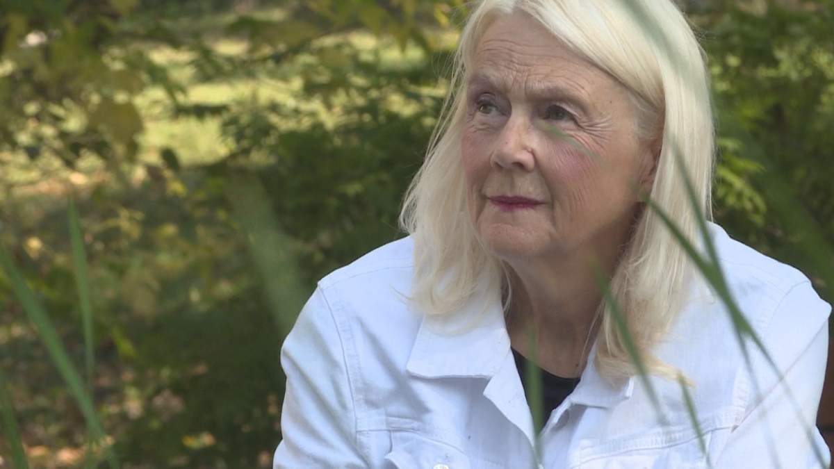 A woman with blonde hair looks up into the distance. She is wearing a white jacket and black top. Blades of grass in the foreground partially cover her face.