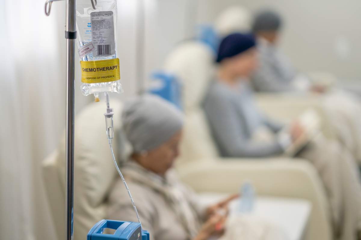 Three women sit in individual chairs as they receive their Chemotherapy by intravenous. They are each dressed comfortably and have head scarves on to keep them warm as they engage in the activity they each brought to keep them occupied.