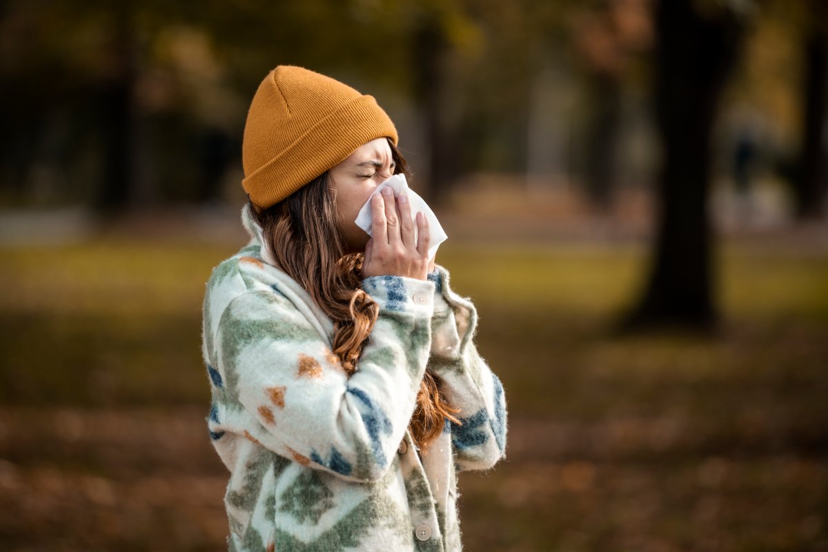 A woman in a beanie holding a tissue to her nose while standing outdoors in autumn.