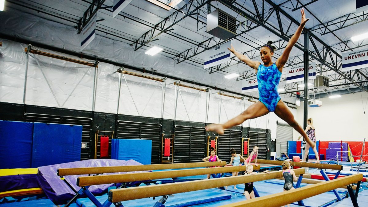 A gymnast leaps on a balance beam in a gym. Her gym suit is blue, and her hair is in a bun,