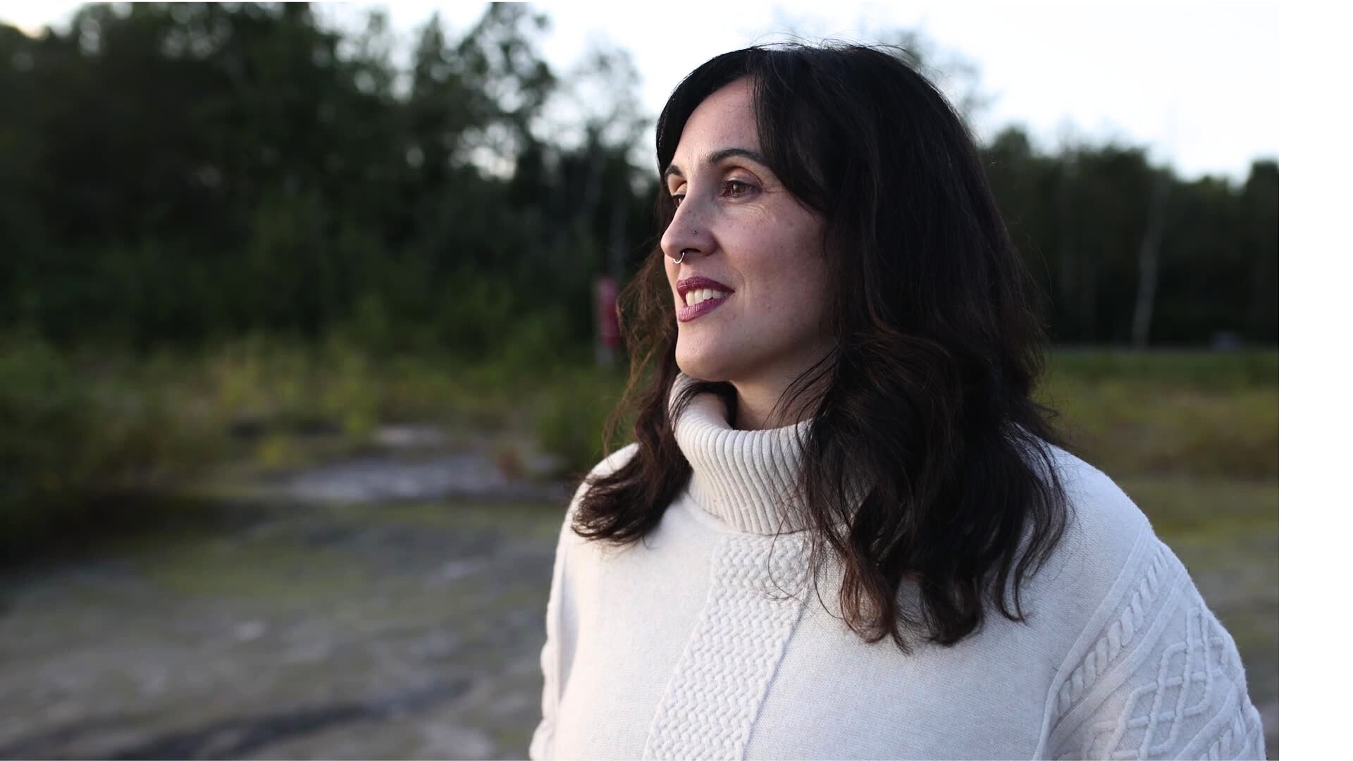 Ashlee Cunsolo is looking towards the sunset wearing a cream coloured knit sweater. Her dark hair is wavy and drapes down her shoulders.