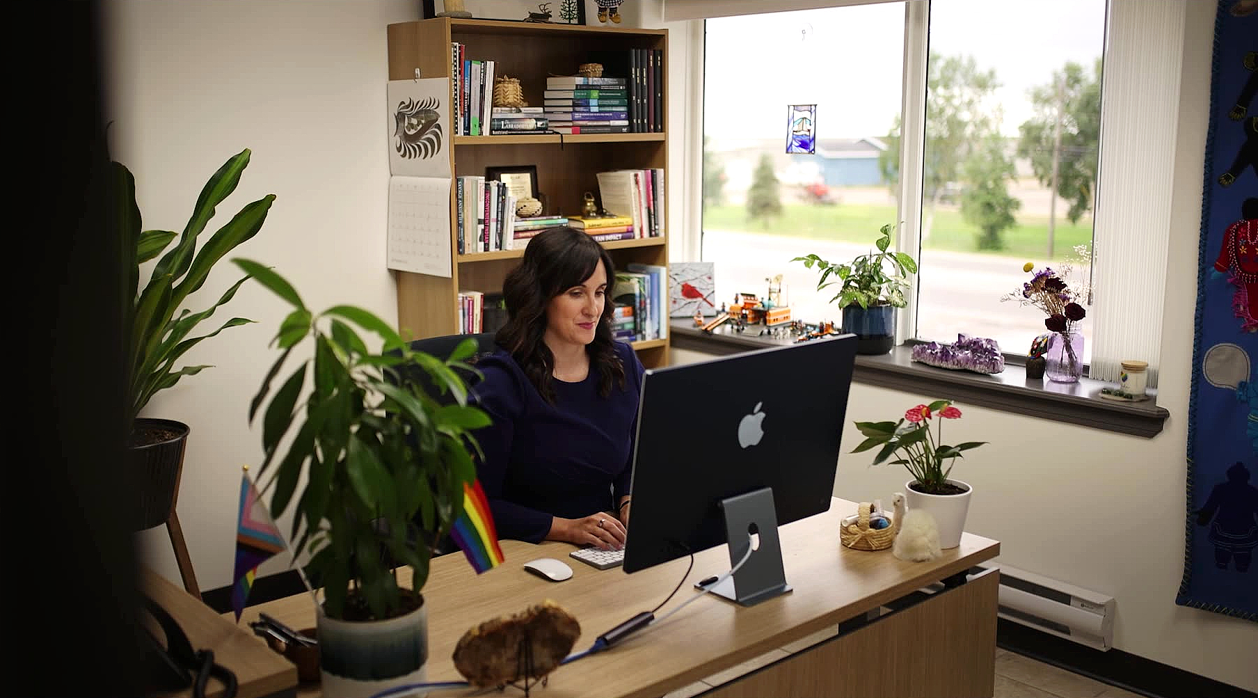 Ashlee Cunsolo is wearing a blue shirt sitting in her office, typing at her computer. Her dark hair is curled and she is surrounded by plants, books, and artwork in her office.