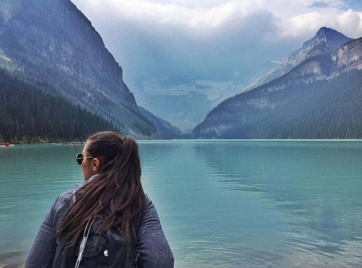 A young woman with brown hair in a ponytail looks out at mountains across a turquoise lake.