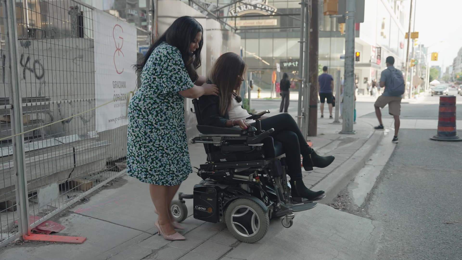 A woman standing on the street helps a woman in a wheelchair descend a steep sidewalk ramp.